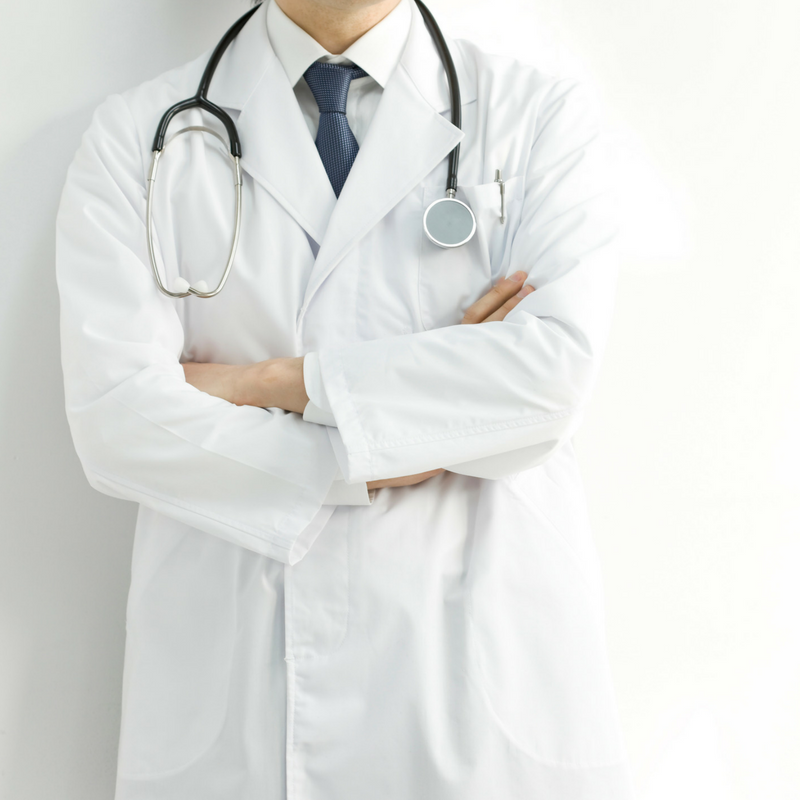 A doctor in a white coat with a stethoscope draped around the neck stands with arms crossed. The background is plain white, and the doctor wears a white shirt and dark tie. The focus is from the neck down.