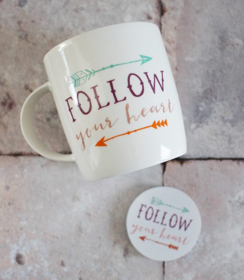 White mug with the phrase Follow your heart in colorful text alongside decorative arrows, placed on a stone surface. Nearby is a round coaster with the same design and text.