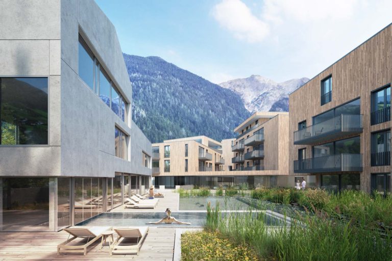 A modern residential complex with wooden and concrete buildings surrounds a serene pool. Lounge chairs line the poolside. In the background, misty mountains rise under a cloudy blue sky. Greenery is interspersed around the pool area.