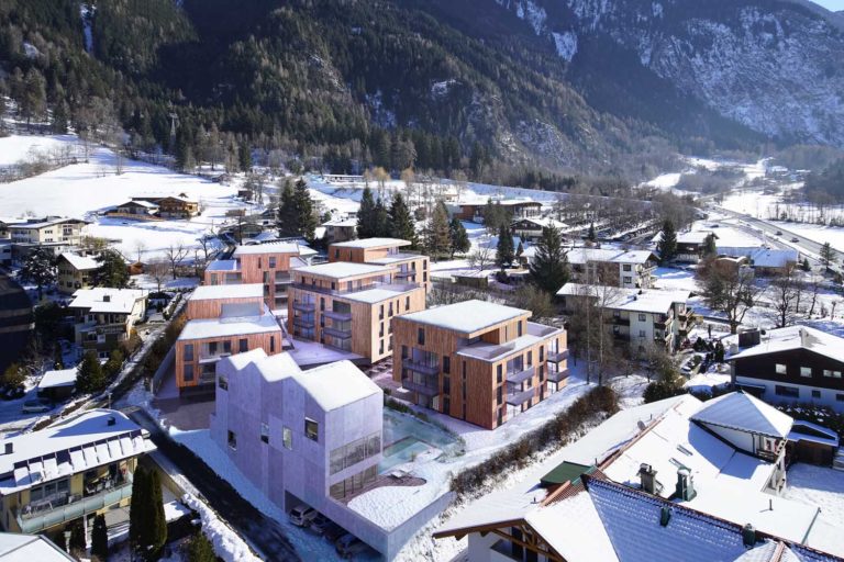 Aerial view of a snowy alpine village, featuring modern multi-story buildings with wooden elements. Snow-covered rooftops and trees surround the structures, set against a backdrop of mountains and forested landscape.