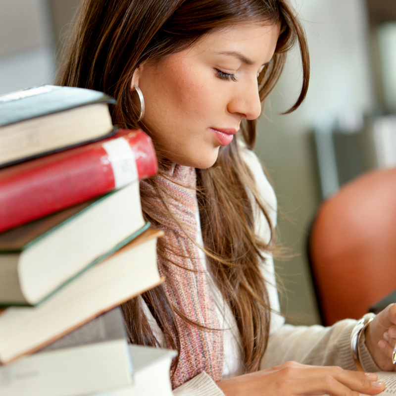 A woman with long brown hair sits at a table, focused on reading a book. She wears hoop earrings and a scarf. A stack of books is piled in front of her, and the background is softly blurred, suggesting a library or study space.