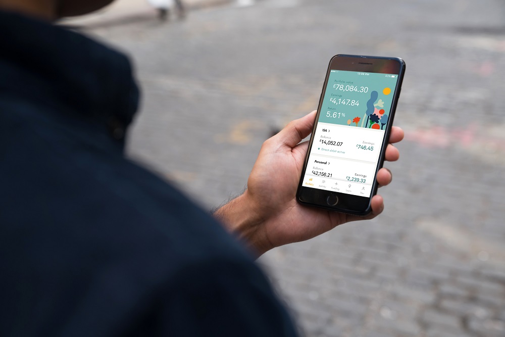 A person holds a smartphone displaying a colorful financial app with graphs and numbers, showing account balances and statistics. The background is a blurred cobblestone street.