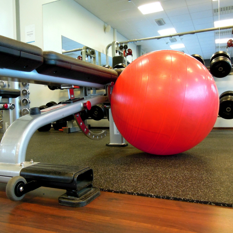A gym scene with a bright red exercise ball next to a weight bench. Various gym equipment, including dumbbells on racks, is visible in the background. The lighting is bright, showcasing a clean and organized workout space.