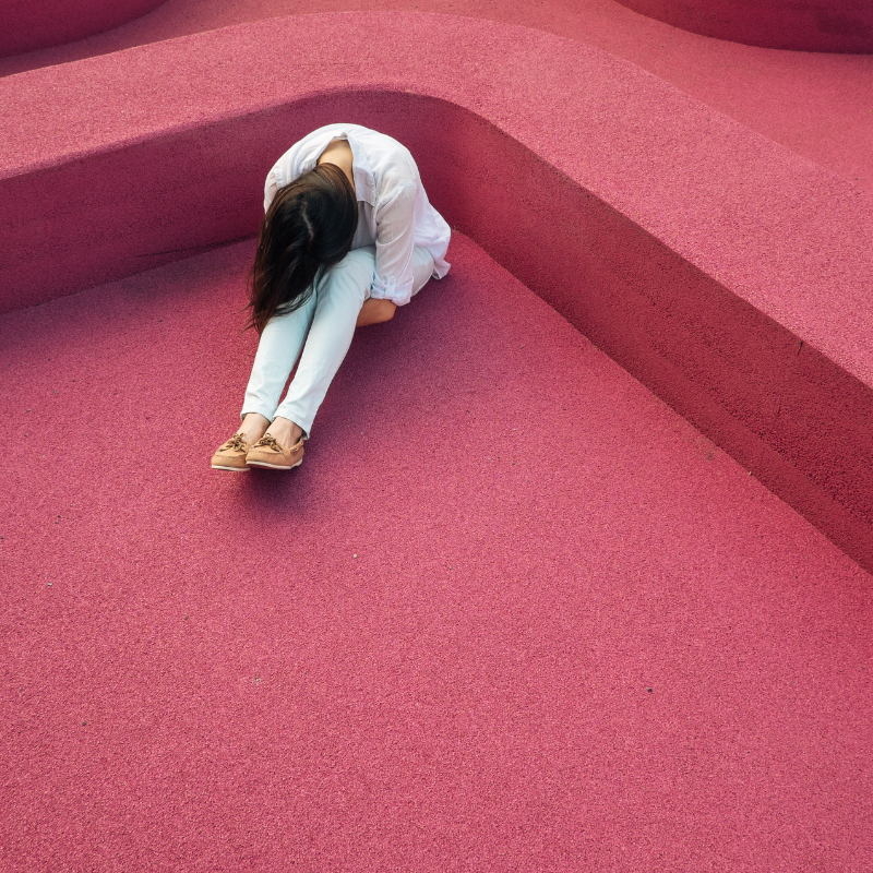 A person in casual clothing sits on a textured pink surface with their head resting on their knees, surrounded by curved pink structures. The scene conveys solitude and introspection.