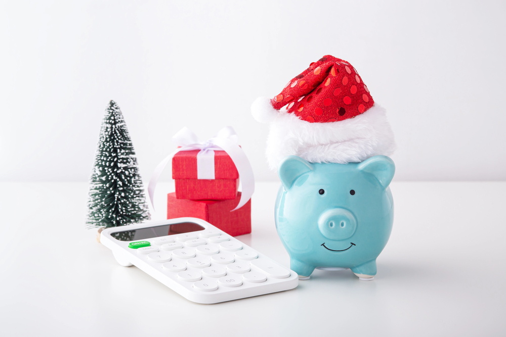 A light blue piggy bank wearing a Santa hat sits next to a calculator, two stacked red gift boxes, and a miniature Christmas tree, serving as a reminder to start saving for Christmas.
