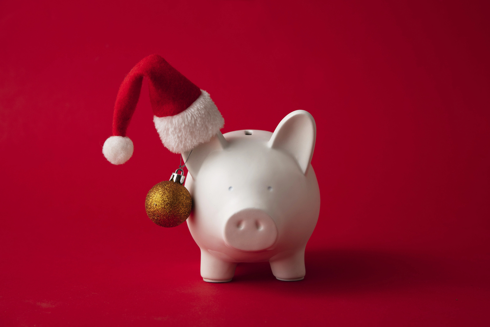 A white piggy bank wearing a red Father Christmas hat and a small gold ornament against a red background, serving as a reminder to start saving for Christmas.