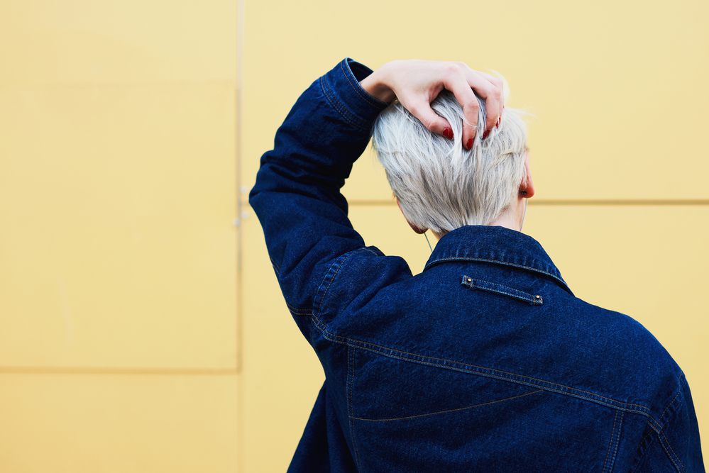 Rear view of female blonde hipster dressed in trendy jeans jacket with copy space area for your brand name or advertising posing on urban setting wall background. Female with stylish haircut posing