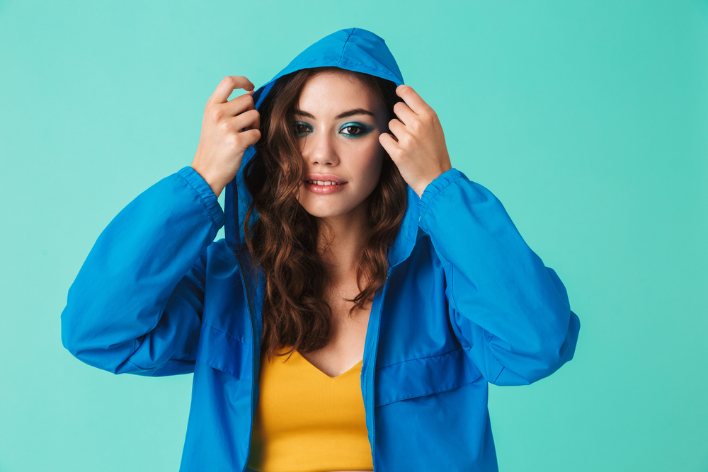 Photo of european young woman 20s in streetwear putting on hood of raincoat or jacket isolated over blue background