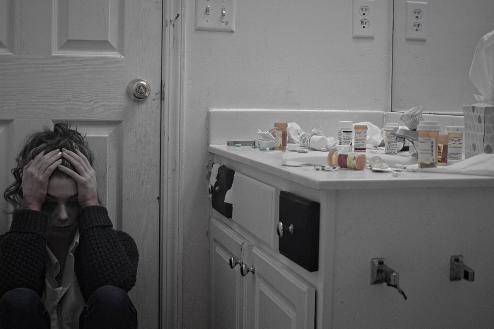 person sat with head in hands in a bathroom with pill bottles on the side