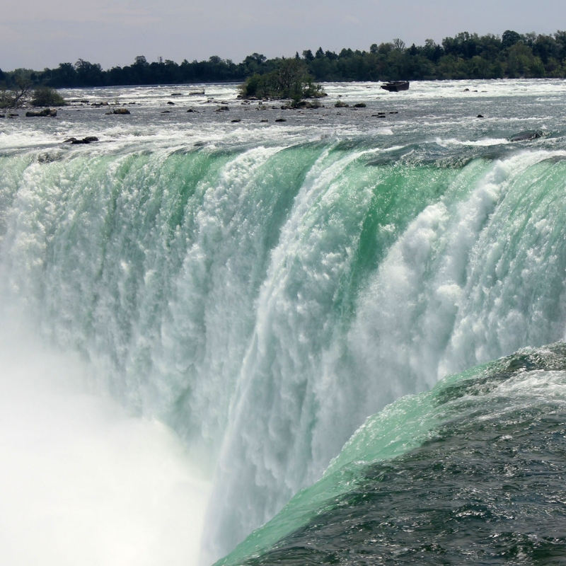 A powerful waterfall cascades over a wide ledge, creating a misty spray below. The water is a vibrant green-blue, and trees line the horizon under a clear sky.