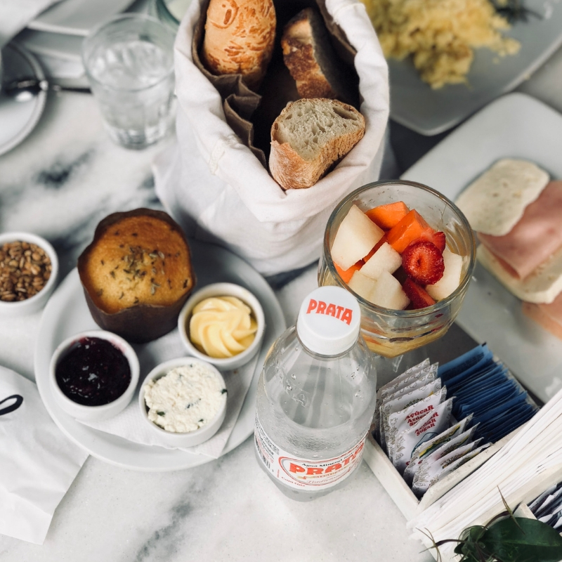 A breakfast spread on a white marble table, including a basket of bread, fruit salad in a glass, a muffin, jam, butter, cream cheese, a bottle of Prata mineral water, and packets of sugar.