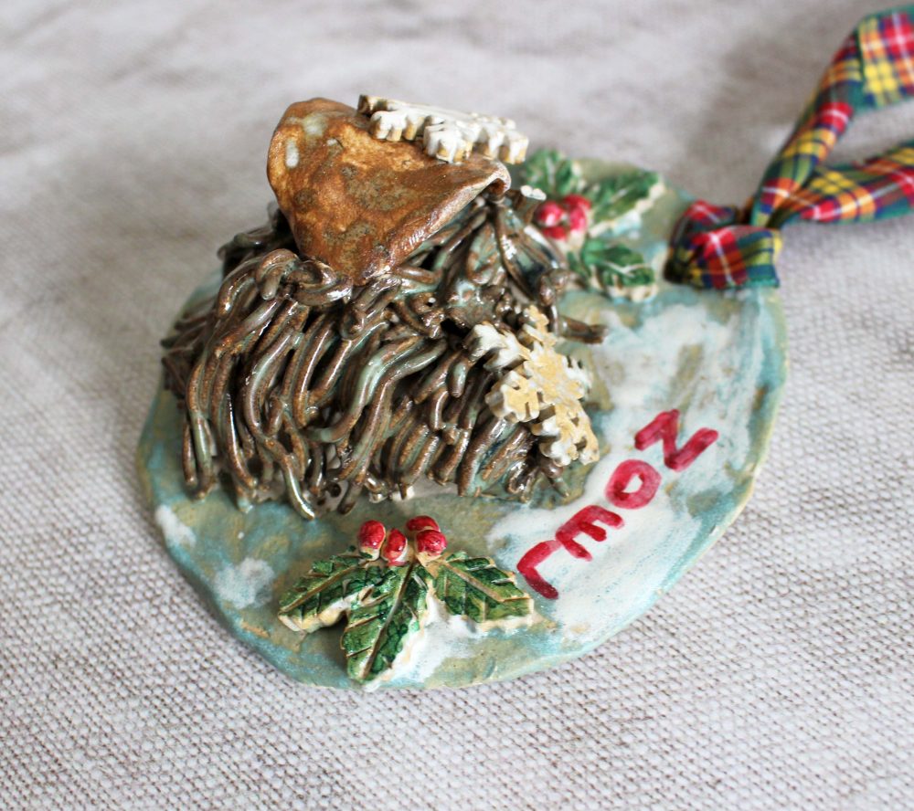 A handmade ceramic ornament by D. H. Potteries features a cozy cottage with a brown roof, surrounded by greenery and holly with red berries. 2007 is inscribed in red, and a plaid ribbon is attached for hanging against a gray background.