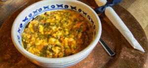 A ceramic bowl filled with a creamy, spinach-based lentil stew sits on a copper plate. A spoon and a napkin tied with a dark ribbon are placed beside it. The stew appears rich with visible chunks and a mix of green and yellowish tones.