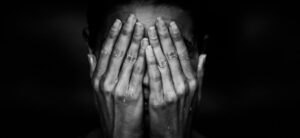 A person covers their face with both hands. The image is in black and white, highlighting water droplets on the hands and fingers. The background is dark, creating a dramatic and emotional atmosphere.