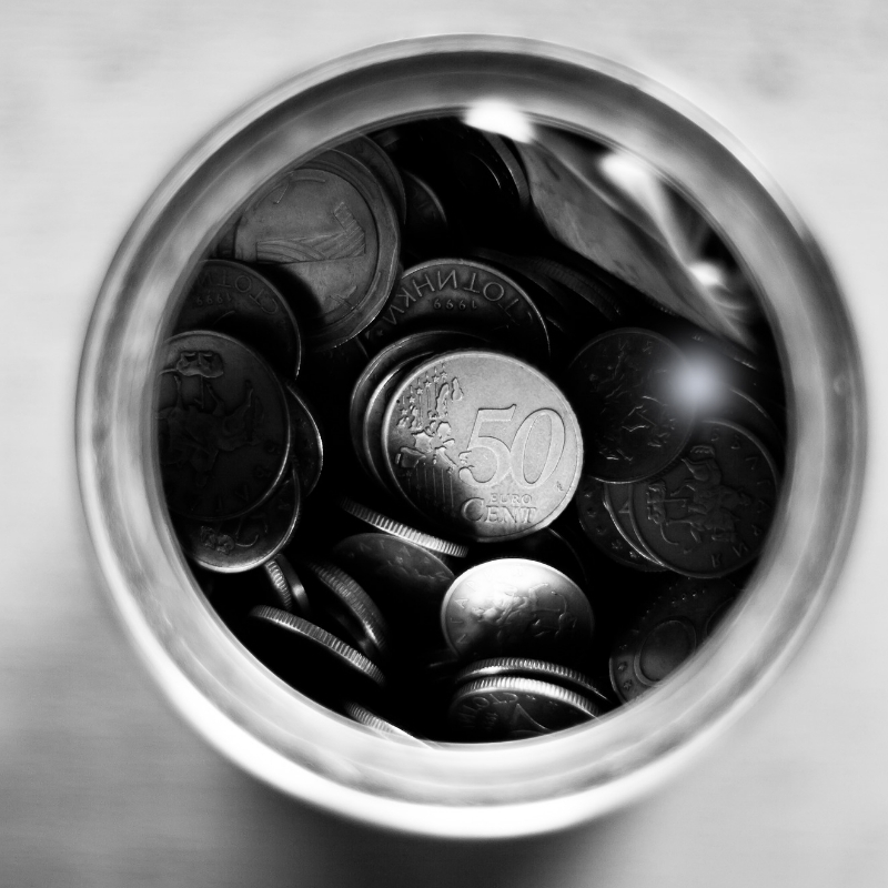 A black-and-white image shows a glass jar filled with assorted coins, including a prominently displayed 50 cent coin—a perfect symbol of the resolve to save money. The jar is viewed from above, capturing the circular shape and shiny metallic surfaces of the coins.