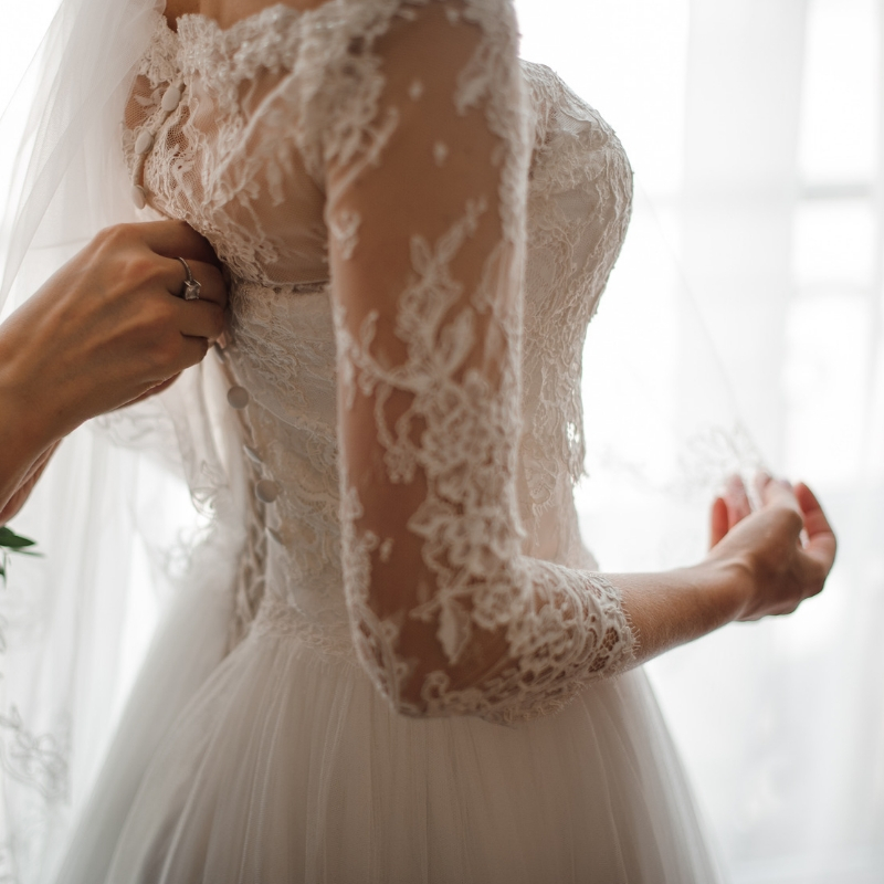 A person wearing a lace wedding dress is having the back of their dress adjusted. Natural light illuminates the intricate lace details of the gown. The persons veil is partially visible.