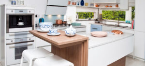 Modern kitchen featuring white Grundig appliances and cabinets. A small wooden breakfast bar with white chairs displays blue and white dishware. Copper pans and decorative items line the shelves, while a window offers a refreshing view of greenery outside.