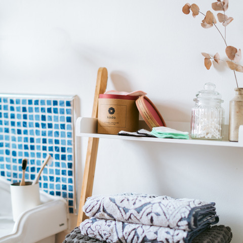 A bathroom shelf with a container, a glass jar of cotton swabs, dried leaves in a vase, and folded towels. Theres a blue mosaic decor in the background.