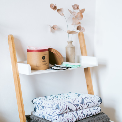 Wooden ladder shelf with a white top holds a brown jar labeled Kado, a small vase with dried leaves, and folded cloths. Below, patterned towels are neatly stacked. The scene is set against a plain light-colored wall.