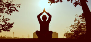 A person sits cross-legged on a surface, silhouetted against the sunrise. They have their hands together above their head, as if meditating or practicing yoga. Trees frame the image, with a cityscape faintly visible in the background.