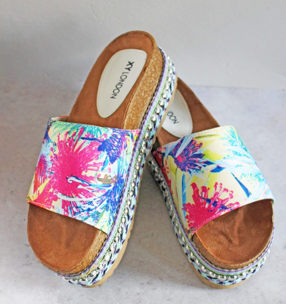 Colorful slide sandals with floral patterns in pink, blue, and yellow. They have a thick sole with a woven design and are labeled XY LONDON on the insoles. They are displayed on a light gray surface.
