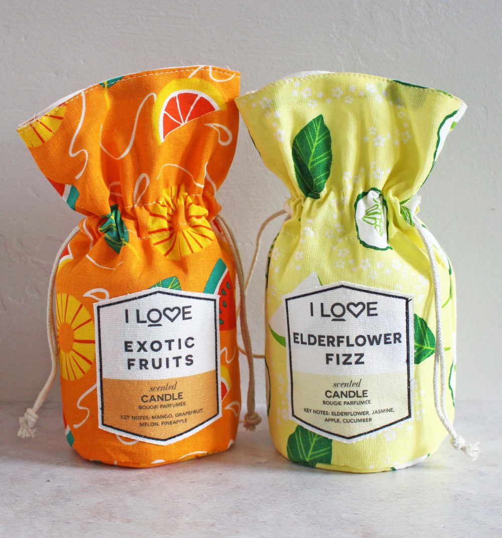 Two decorative drawstring pouches with printed fruit patterns. The orange pouch is labeled Exotic Fruits and the yellow pouch Elderflower Fizz. Each contains a scented candle, with key notes listed on the label.