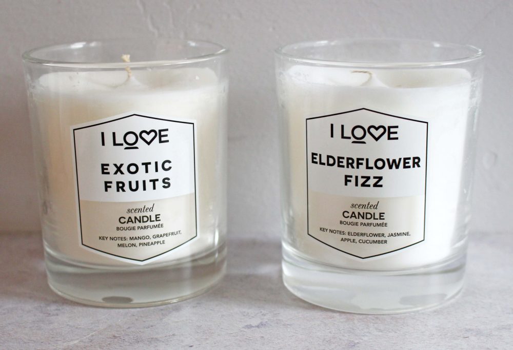 Two glass candles with white wax are labeled Exotic Fruits and Elderflower Fizz. The Exotic Fruits candle has notes of mango, grapefruit, melon, pineapple. The Elderflower Fizz candle has notes of elderflower, jasmine, apple, cucumber.