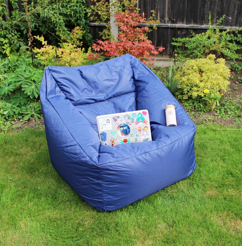 A blue bean bag chair on green grass in a garden, surrounded by leafy plants. A laptop with colorful stickers and a cylinder-shaped drink container rest on the chair.