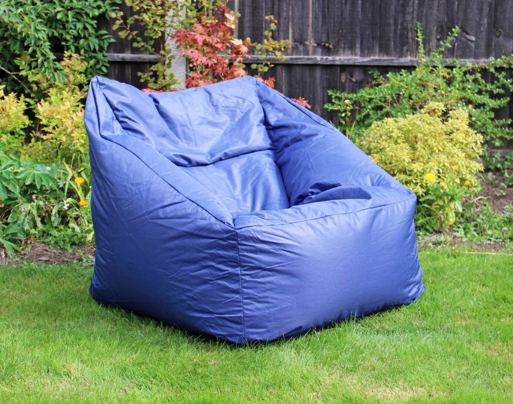 A large blue bean bag chair sits on a green lawn. Surrounding it are various plants and shrubbery in a garden setting, with a wooden fence in the background.