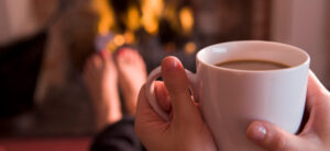 Close-up of a person holding a white mug filled with coffee or tea. The background features a cozy fireplace with flames burning, and a pair of feet with red-painted toenails resting nearby. The scene suggests warmth and relaxation.