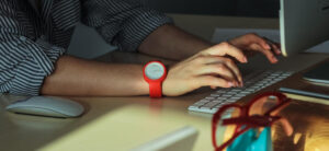 Person wearing a red smartwatch types on a keyboard at a desk. A computer mouse is nearby, along with a pair of red glasses and a turquoise object. Sunlight casts shadows across the scene.