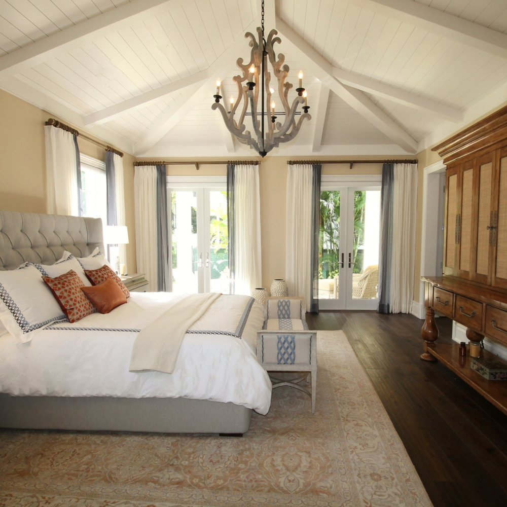 A spacious bedroom with a vaulted ceiling and chandelier. The room features a large bed with white and blue bedding, wooden furniture, and a sliding door opening to a garden view. Soft natural light fills the space, highlighting a patterned rug.