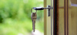 Close-up of a door slightly ajar with a set of keys in the lock. The keys have a black plastic head, and the background is a blurred outdoor scene of greenery.