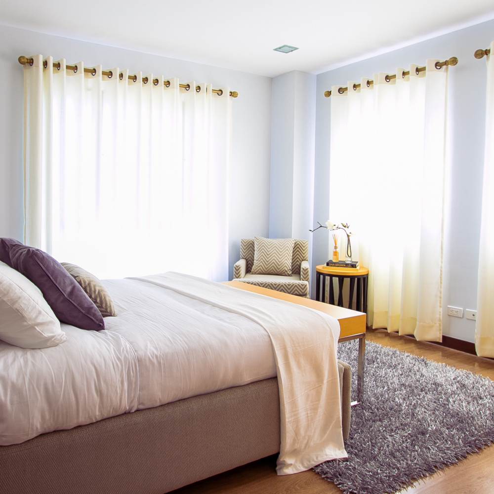 A bright bedroom with a large bed, white pillows, and a beige blanket. Sheer curtains allow natural light through two windows. A gray rug covers part of the wooden floor. A small table and armchair are near the window.