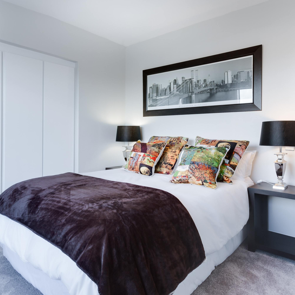 A cozy bedroom with a neatly made bed featuring a dark brown throw and colorful accent pillows. Two bedside tables with lamps flank the bed. A framed black-and-white cityscape photo hangs above the bed. The room has a tranquil ambiance.