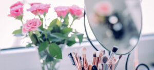 A vanity table with various makeup brushes in a holder, reflected in an oval mirror. A bouquet of pink roses with green leaves is placed nearby, softly out of focus. Bright natural lighting enhances the scene.