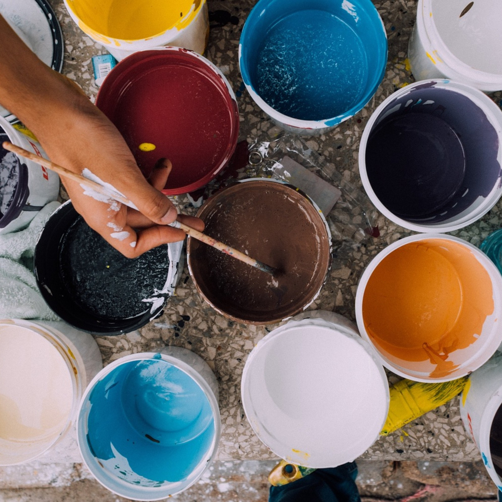A hand holds a brush over a container of brown paint amid several open paint buckets of various colors, including red, blue, black, white, and yellow. The buckets are placed on a speckled floor, surrounded by paint splatters.