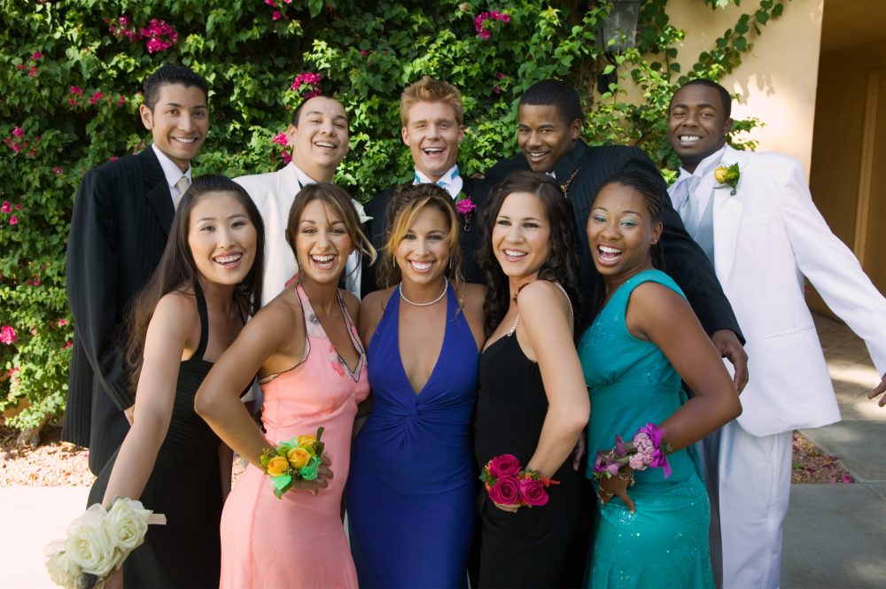 A group of eight people at a formal event, with women in colorful dresses and men in suits. They are smiling and standing outdoors in front of green foliage. Some are holding flower corsages.