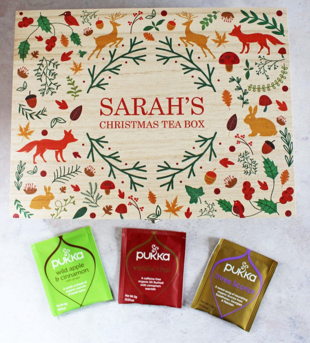 A festive tea box labeled Sarahs Christmas Tea Box is decorated with holiday illustrations of reindeer, birds, and leaves. Below the box, there are three Pukka tea packets: Wild Apple & Cinnamon, Vanilla Chai, and Three Licorice.