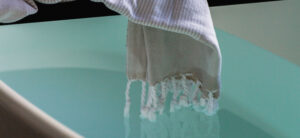 A light gray towel with fringed edges is draped over the edge of a bathtub, dipping slightly into the pale turquoise water inside. The scene conveys a serene and inviting atmosphere.