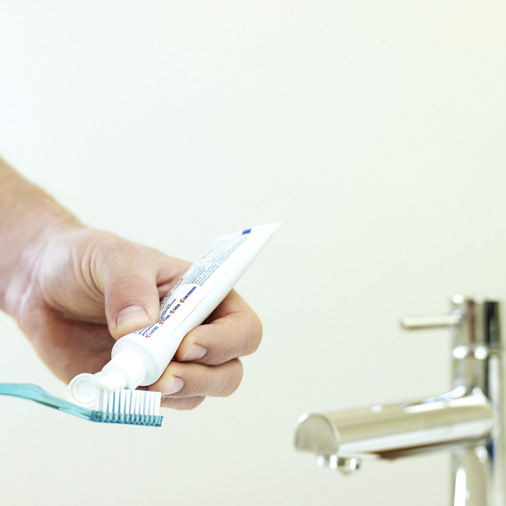 A person holding a tube of toothpaste, squeezing it onto a blue toothbrush. A metallic faucet is visible nearby, all set against a plain light-colored background.