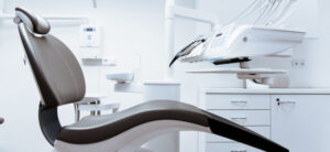 A clean, modern dental clinic with an empty dentist chair, overhead dental equipment, and white cabinetry. The scene is well-lit and organized, conveying a sense of hygiene and readiness for a dental procedure.