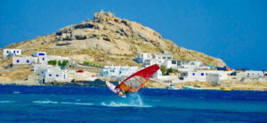 A windsurfer with a bright sail glides across blue sea waves in front of a rocky island with white buildings. The island is under a clear, blue sky, and small boats are anchored near the shore.