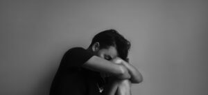 A person in a black shirt sitting against a wall, with their head resting on their knees and arms wrapped around their legs, conveying a sense of sadness or contemplation. The image is in black and white.