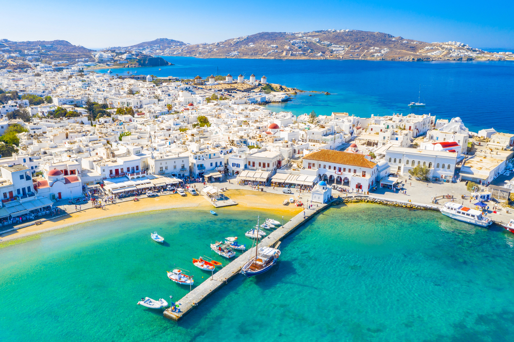 Aerial view of Mykonos town with white buildings along the coastline, a pier extending into turquoise water, and several small boats anchored in the harbour.