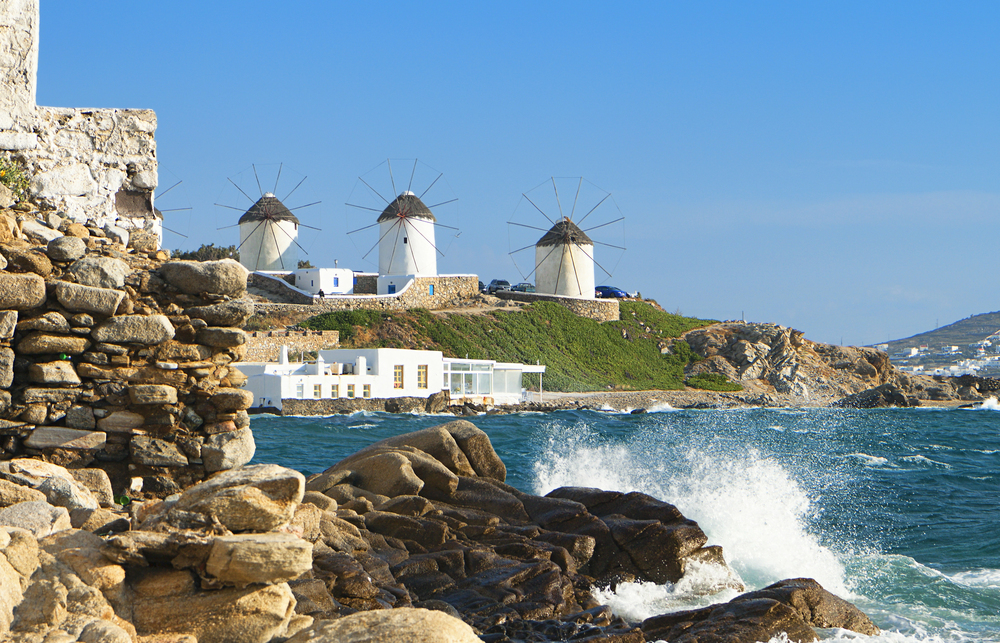 Three traditional white windmills on a hill overlooking the sea, with waves crashing against rocks in the foreground.