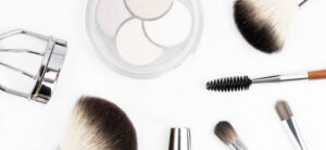 A flat lay of makeup tools on a white background, including fluffy brushes, an eyelash curler, a mascara wand, and a palette of neutral eye shadows.