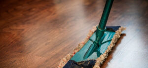 A close-up of a green dust mop cleaning a wooden floor. The mop has a rectangular head covered with a fabric pad, which has a frayed, beige edge. The floor appears clean and polished.