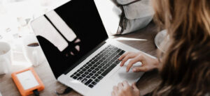 A person typing on a laptop at a wooden desk with a portable hard drive to the left and a gray bag nearby. Sunlight streams in, and two coffee cups are visible, one white and empty, the other filled with dark liquid.