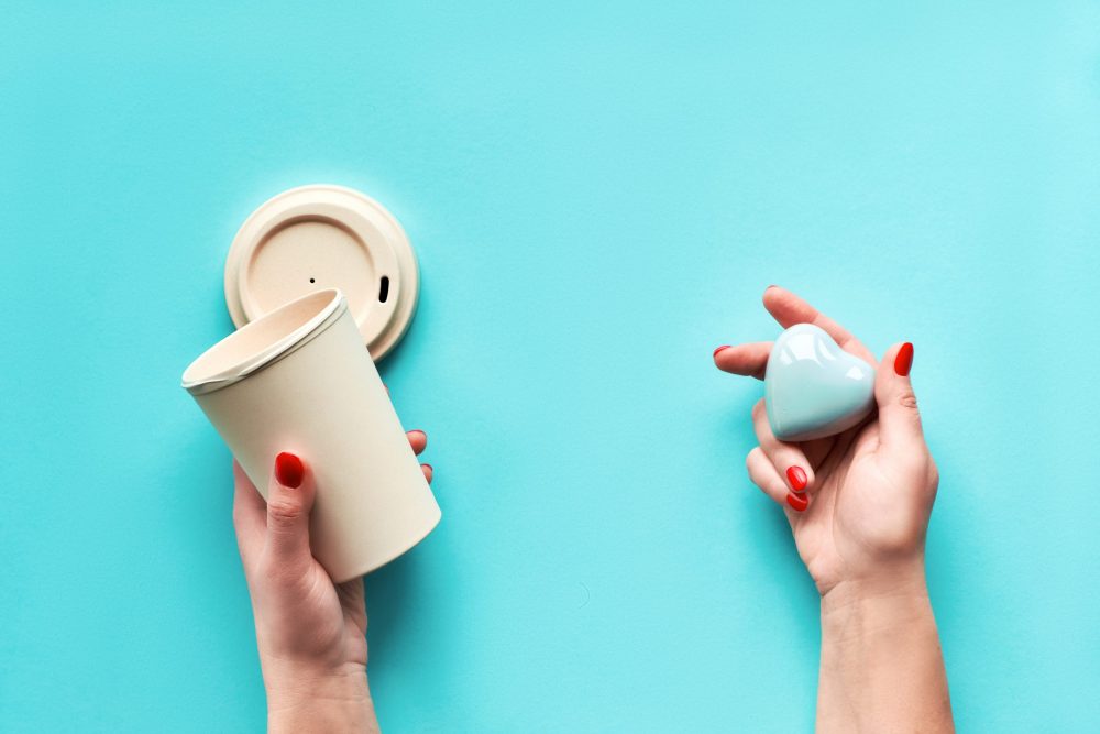 Stylish reusable eco coffee travel mug, bamboo cup with lid and ceramic heart in female hands. Blue mint aqua menthe paper background. Zero waste eco friendly lifestyle trend.